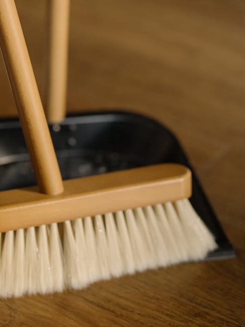 A close-up view of a broom with a wooden handle and beige synthetic bristles, resting inside a black dustpan on a wooden floor. The floor appears clean and well-maintained, highlighting the importance