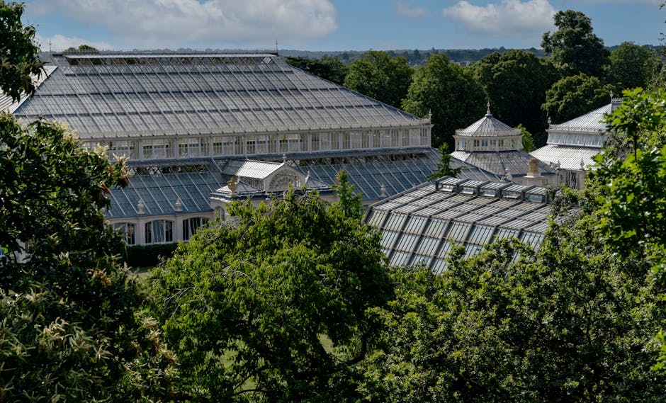 A large Victorian-style glasshouse at Kew Gardens, featuring multiple interconnected structures with glass panels and metal frameworks. The glasshouses are surrounded by lush green trees and foliage,